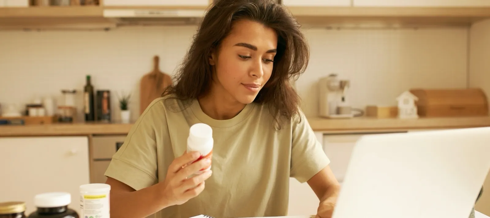Mulher lendo o rótulo de um frasco à mesa da cozinha, com notebook, caderno e outros frascos